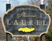Exterior black and beige sign that says welcome to historic laurel, detailed with a line of yellow flowers